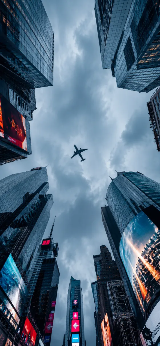 Passenger airplane flying between tall downtown skyscrapers beneath a cloudy blue-gray sky.