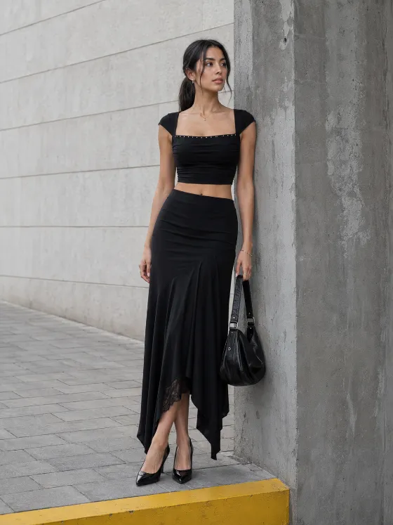 Woman in a black cropped top and long asymmetric skirt standing beside a weathered gray concrete column with a black handbag.