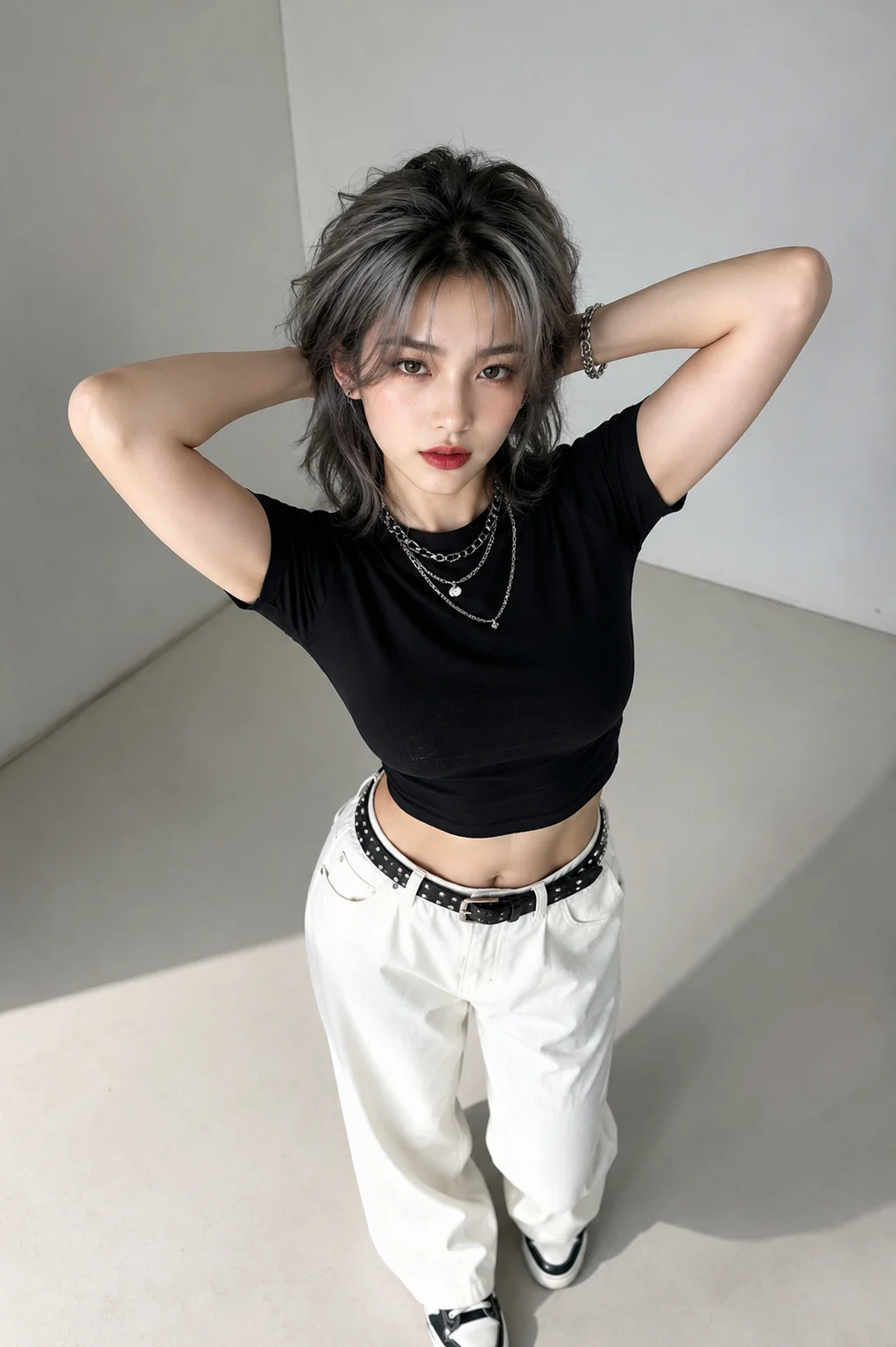 Young woman in a black cropped top and white pants posing with arms behind her head in a gray studio corner.