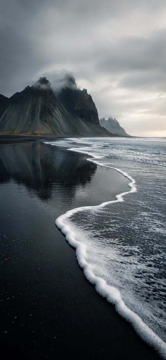 White sea foam curving across a black sand beach toward dark misty mountains under a pale gray sky.