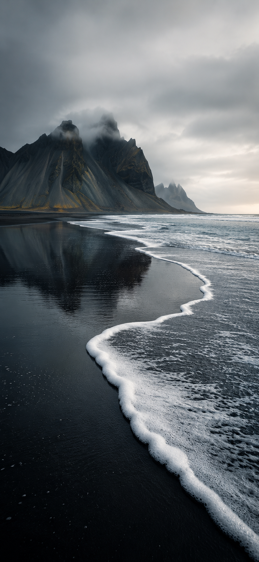 White sea foam curving across a black sand beach toward dark misty mountains under a pale gray sky.