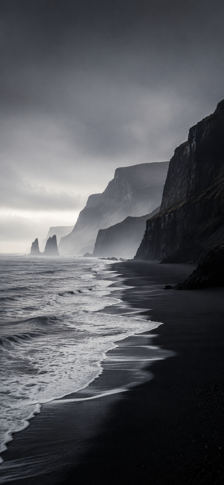 Dark monochrome coastal wallpaper with black sand, white surf, towering cliffs, and misty distant shoreline in a tall iPhone layout.