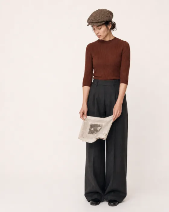 Full-length brown and charcoal fashion look with ribbed knit top, wide-leg trousers, herringbone cap, black shoes, and folded newspaper.