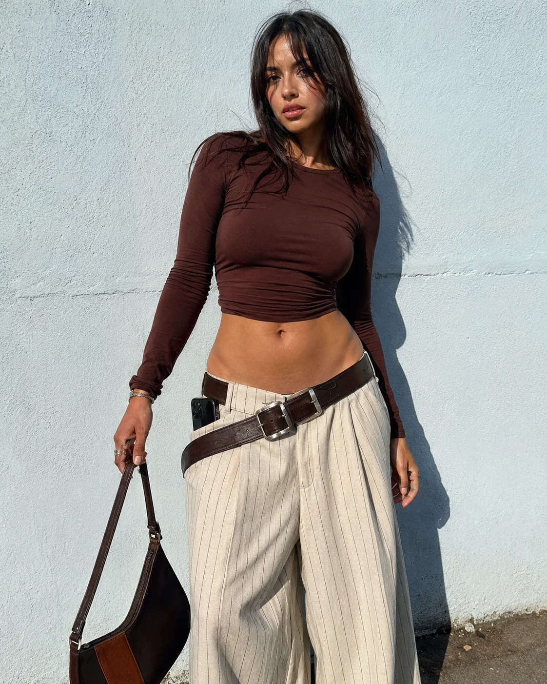 Sunlit Y2K outfit with fitted burgundy crop top, low-rise cream pinstripe trousers, diagonal leather belt, and dark shoulder bag.