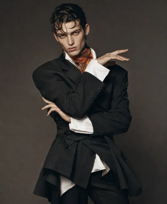 Adult male model in oversized charcoal tailoring and a burnt-orange scarf poses with sculptural hands against a warm-gray studio backdrop.