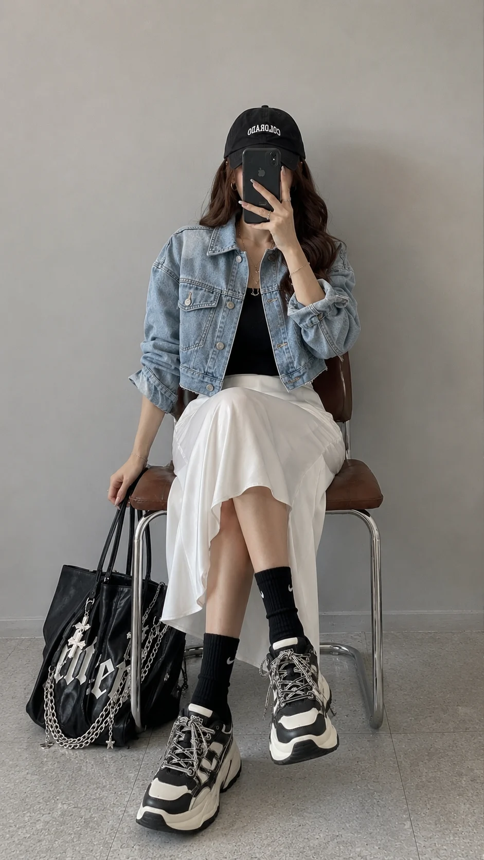 Seated fashion subject in a cropped denim jacket and long white skirt holding a phone in front of the face beside a black tote.