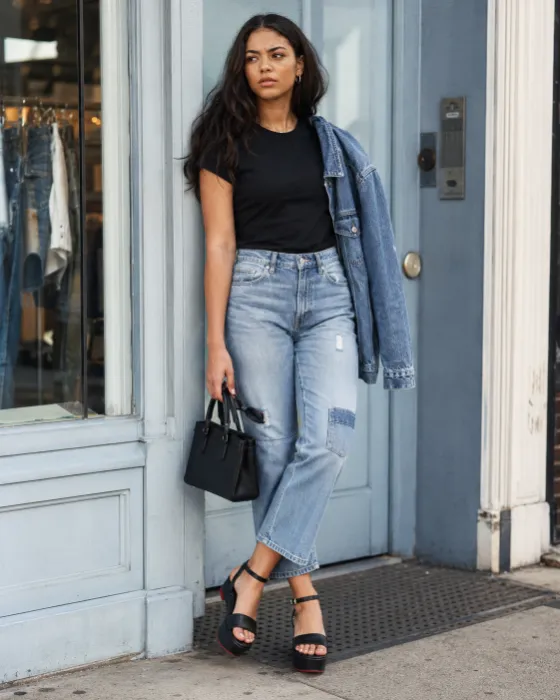 Full-body streetwear portrait with a black tee, light-wash cropped denim, draped jacket, black handbag and pale blue storefront.