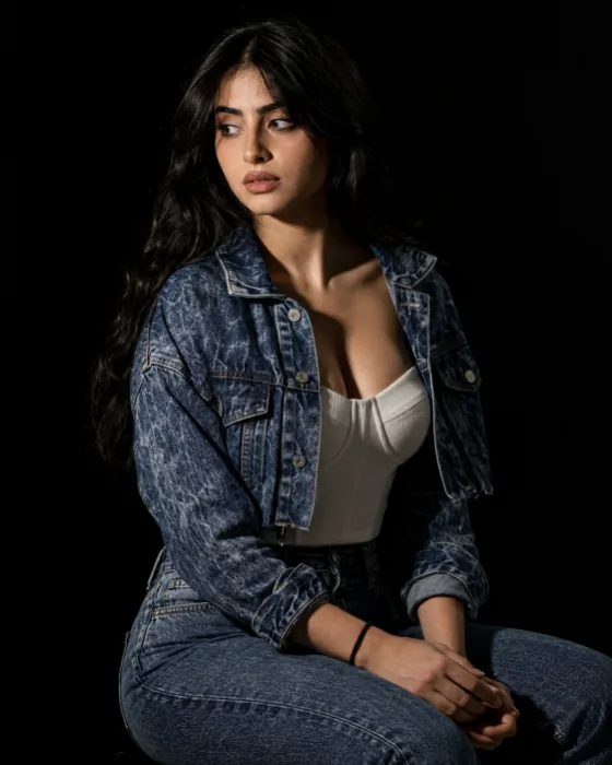 Woman seated in a deep indigo acid-wash denim set with an ivory top, lit by a narrow spotlight against a black background.
