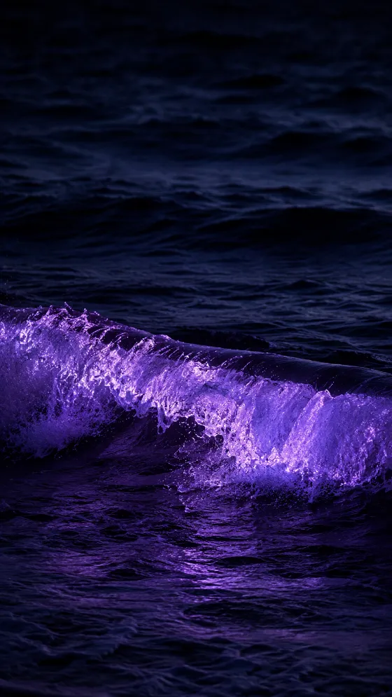 Dark ocean waves at night with vivid violet light illuminating the crest and foam of a breaking wave.