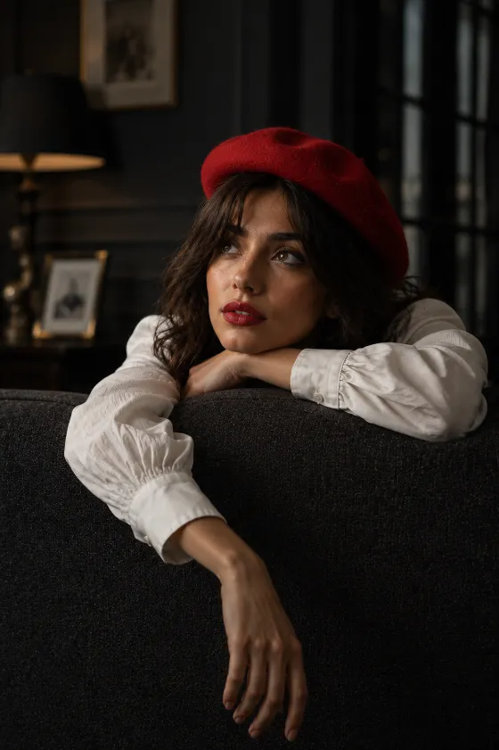 Editorial indoor portrait of a fashion subject wearing a crimson beret and white blouse while leaning over a modern sofa in soft cinematic light.