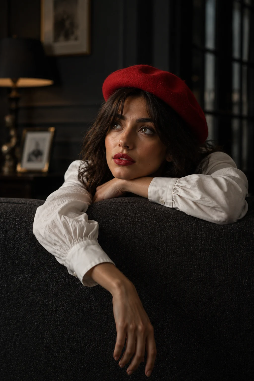 Editorial indoor portrait of a fashion subject wearing a crimson beret and white blouse while leaning over a modern sofa in soft cinematic light.