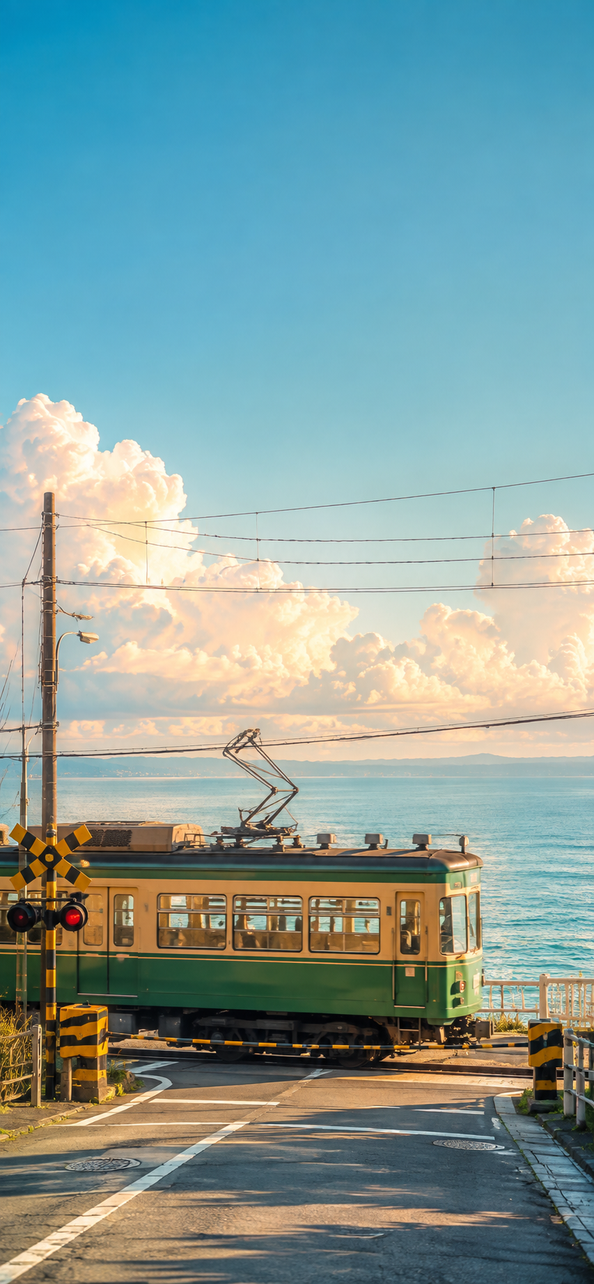 Vintage green electric train passing through a seaside railway crossing beneath a bright blue sky, large clouds, utility poles, and a distant ocean.