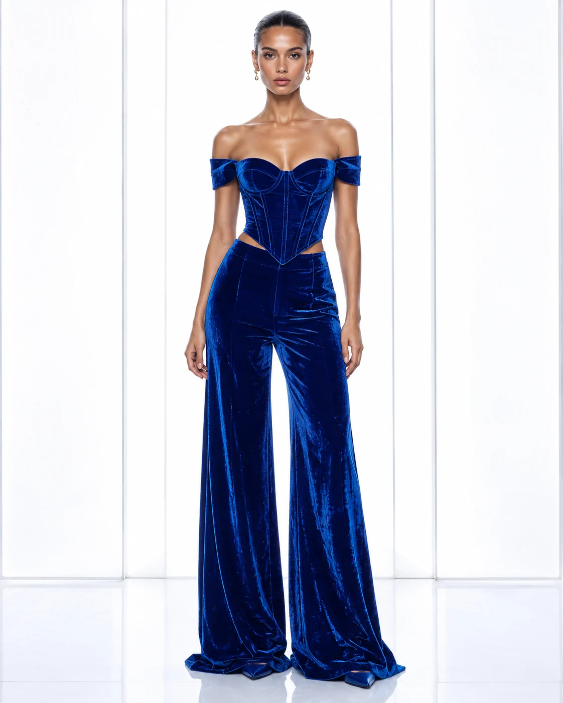 Full-body model in a cobalt blue velvet off-shoulder corset and matching wide-leg trousers in a white studio.