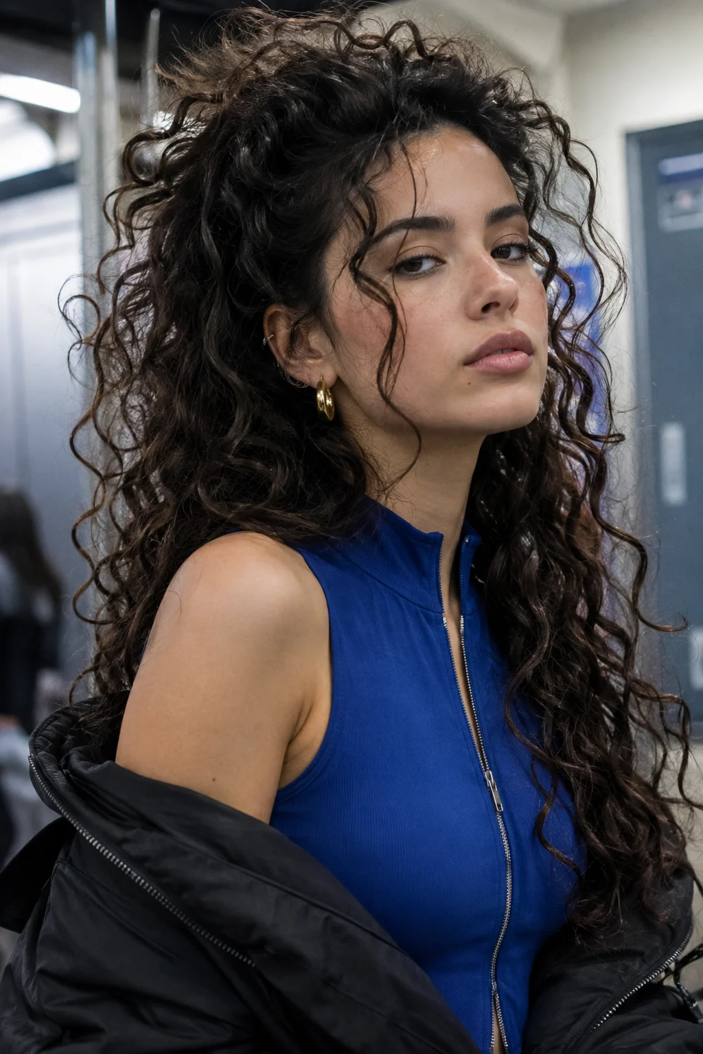Woman with voluminous dark half-up curls wearing a cobalt-blue zip-front top, gold hoop earrings, and a black jacket draped from her shoulders.