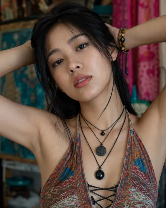 Vertical bohemian fashion portrait with raised arms, layered cord necklaces, loose black hair, and a vivid patterned halter top.