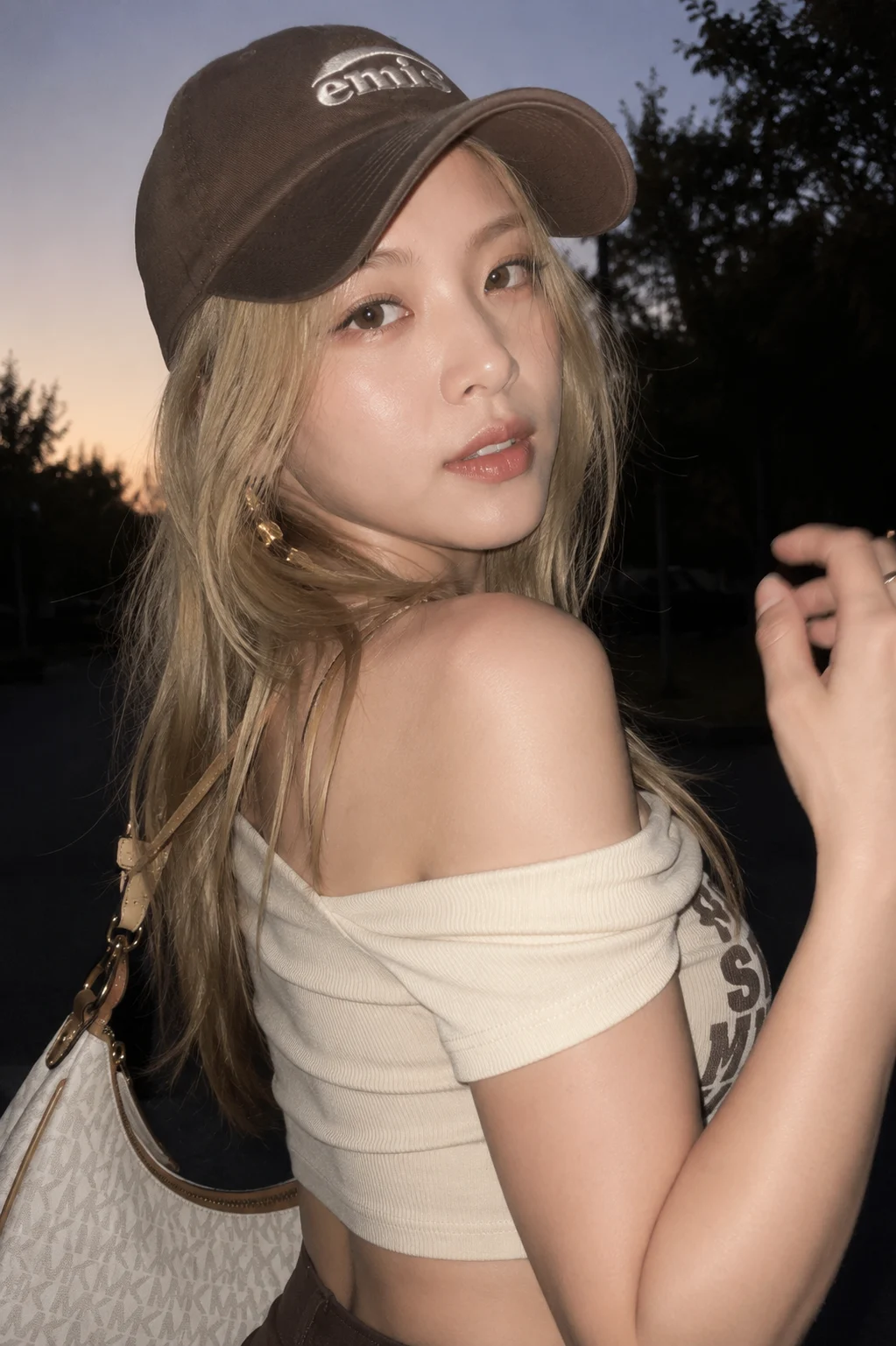 Young woman in a dark baseball cap and off-shoulder light top posing outdoors at dusk with a white shoulder bag.