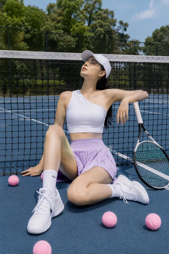 Fashion subject seated on a tennis court beside a racket with pink tennis balls, wearing white athletic top and lavender skirt, leaning against the net.