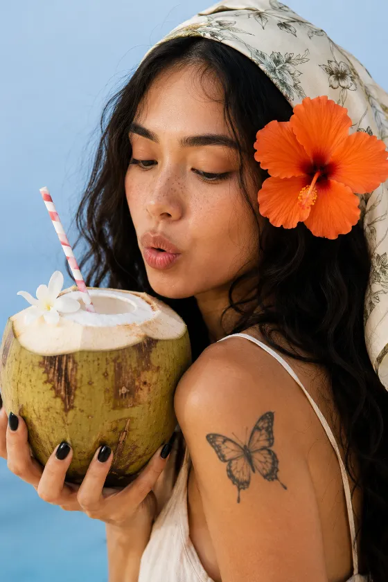 Close-up editorial portrait of a fashion subject holding a fresh coconut with an orange hibiscus flower and botanical headscarf.
