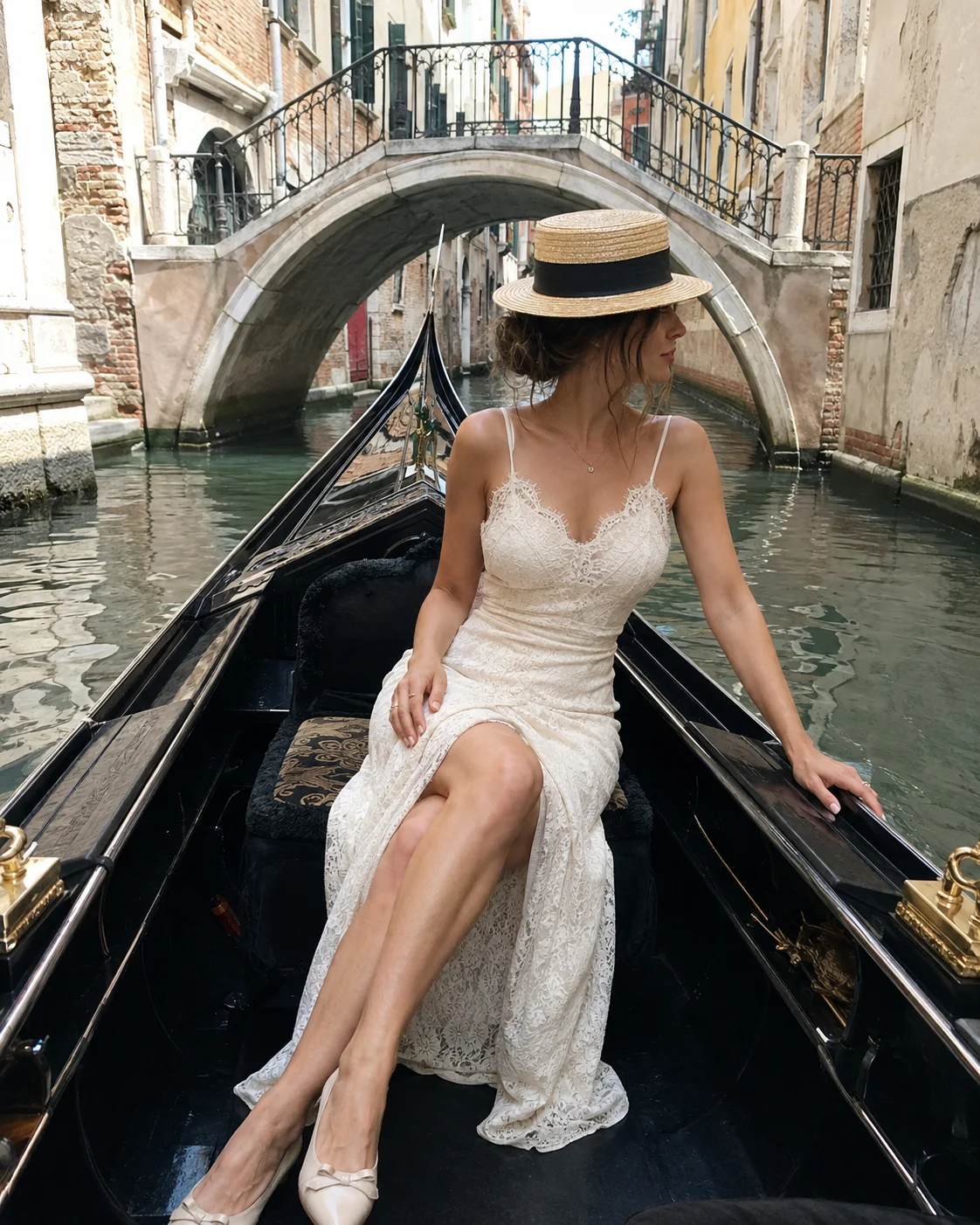 Woman in an ivory lace dress and straw boater hat sits with crossed legs inside a black gondola on a sunlit historic canal.