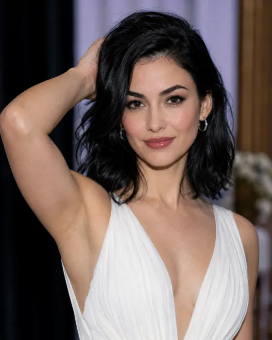 Woman in a white gathered evening gown posing with one arm raised behind her black wavy hair against a softly blurred indoor event background.