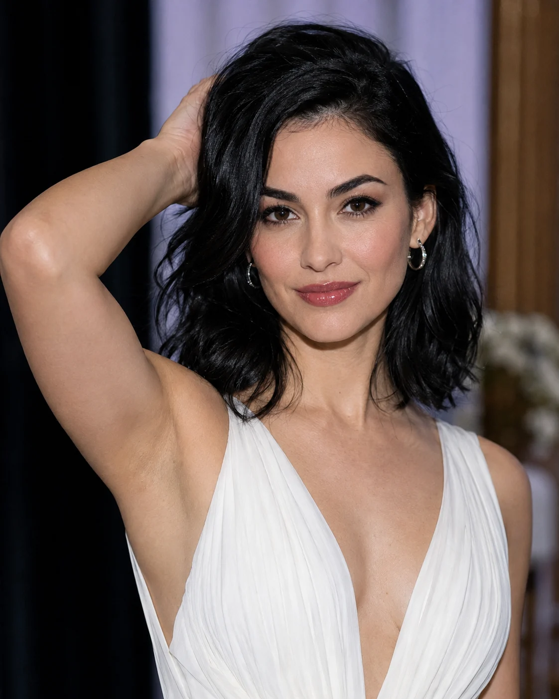 Woman in a white gathered evening gown posing with one arm raised behind her black wavy hair against a softly blurred indoor event background.