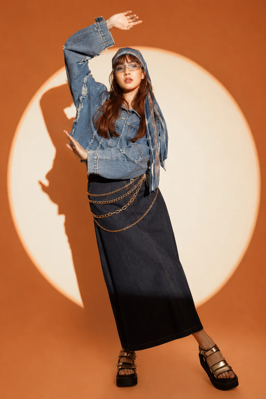 Woman with long auburn-brown hair poses in a distressed denim jacket, long chain-covered denim skirt, visor glasses, and platform sandals.