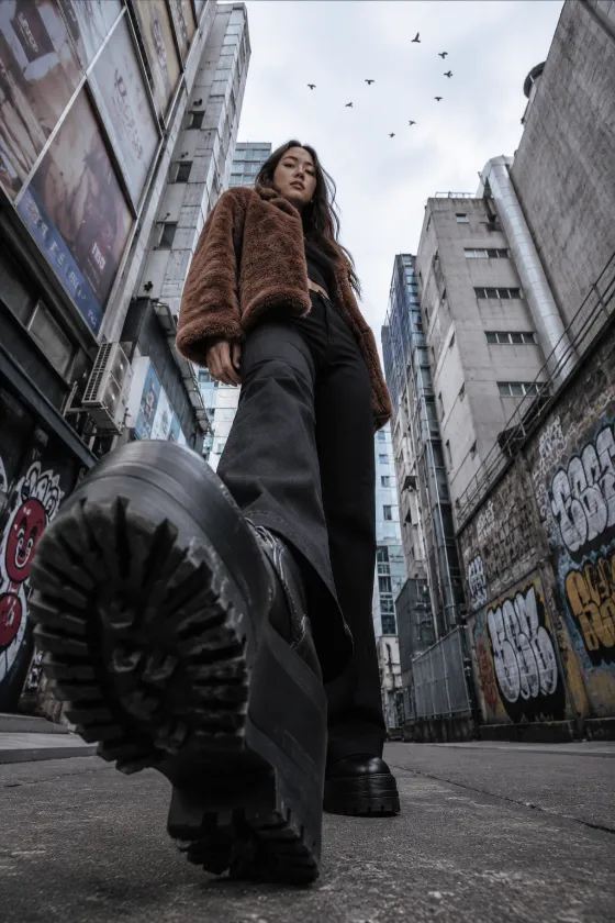Fashion subject walking toward a ground-level camera with a large platform boot dominating the foreground in a graffiti-covered alley.