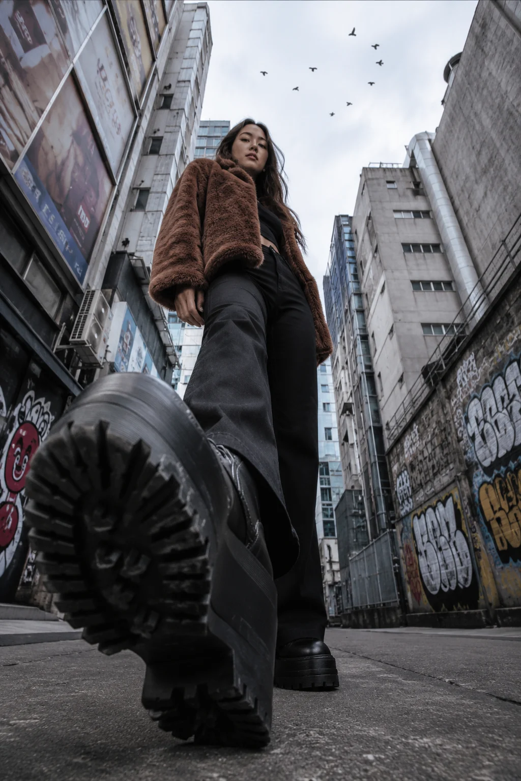 Fashion subject walking toward a ground-level camera with a large platform boot dominating the foreground in a graffiti-covered alley.