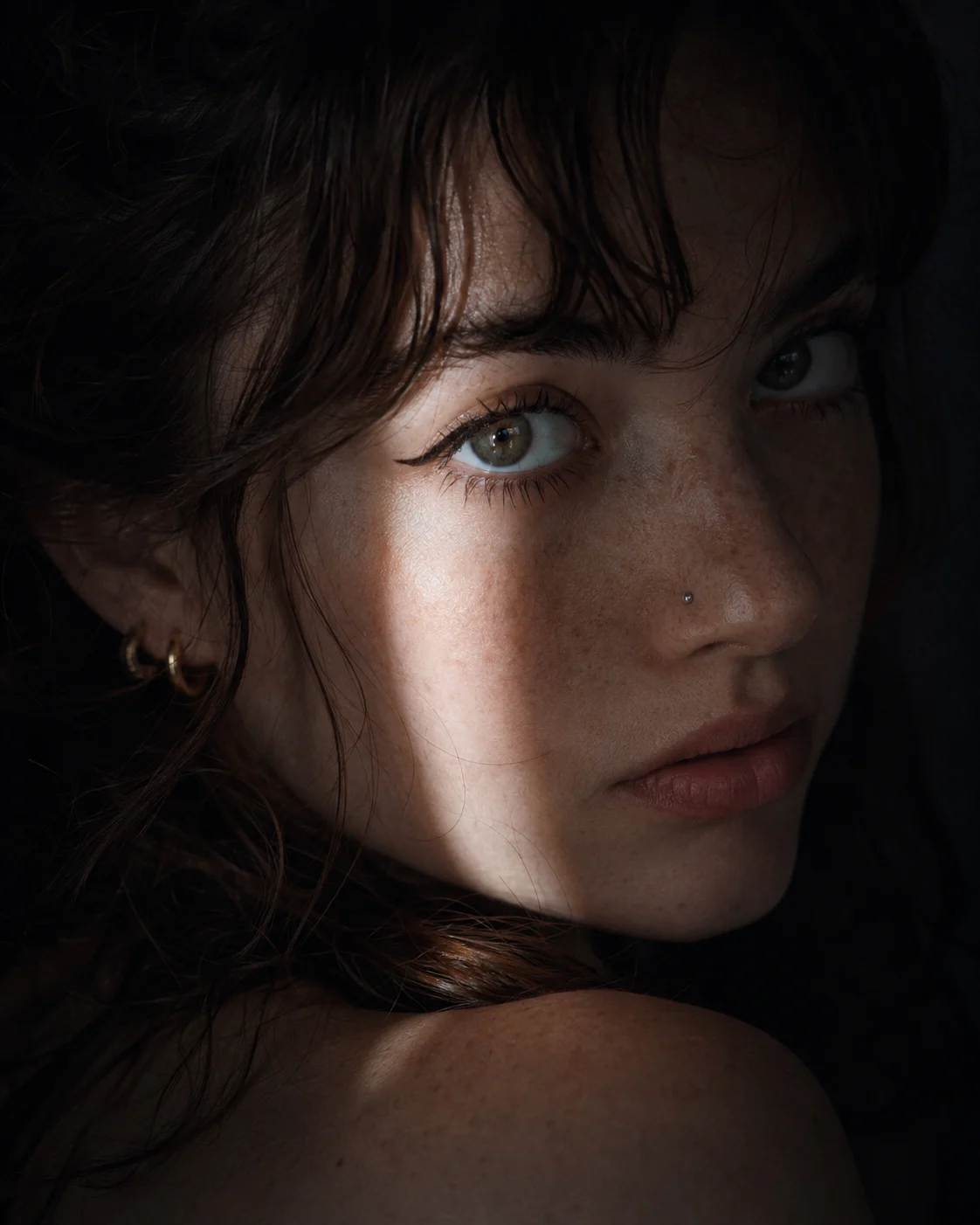 Close-up freckled portrait with dark hair, nose stud, hoop earrings, and dramatic low-key face lighting.