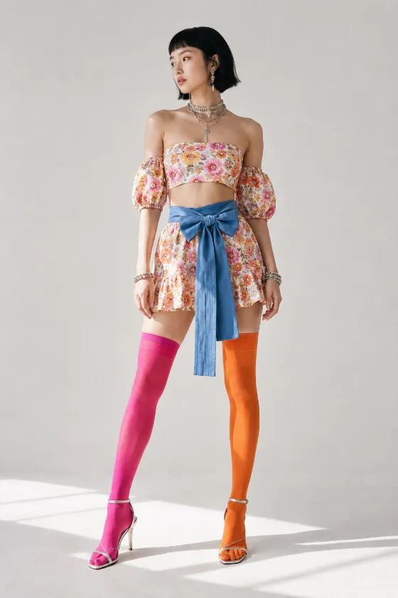 Full-body studio fashion portrait with floral separates, a wide blue bow, pink and orange thigh-high stockings, layered jewelry, and metallic strappy heels.