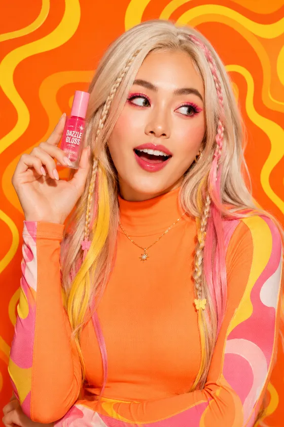 Studio beauty campaign with vibrant orange styling, pastel-highlighted hair, and featured lip gloss product.