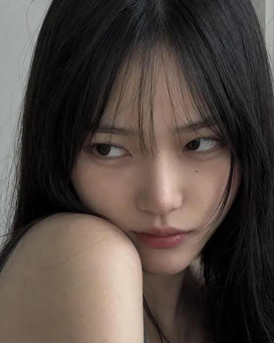 Woman in a tight vertical close-up looking sideways beneath wispy bangs, with long black hair, muted rose lips and a pale cool-gray background.