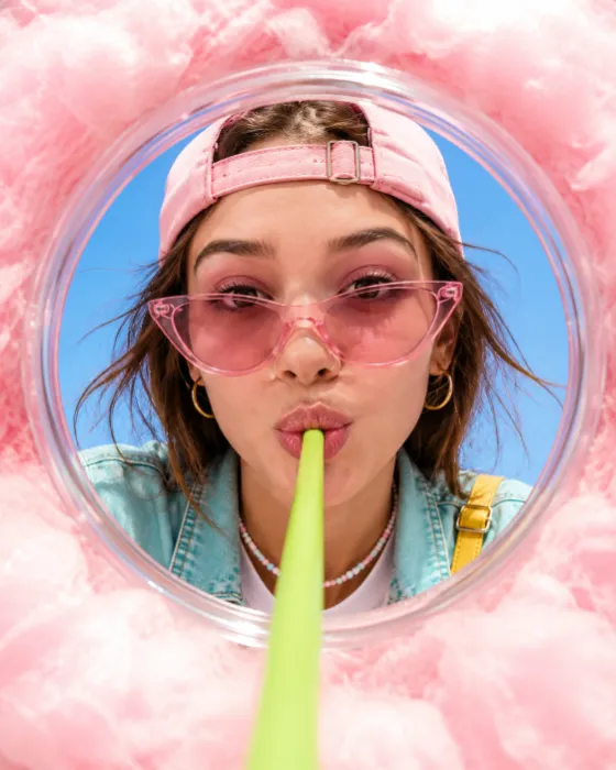 Close-up editorial portrait of a fashion subject framed by pink cotton candy while sipping through a green beneath a blue sky.