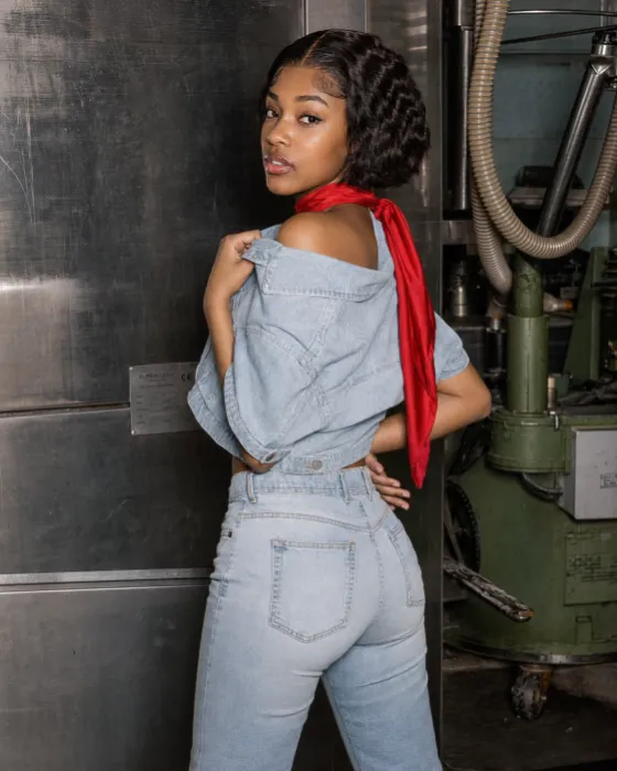 Woman in a cropped light-wash denim jacket and high-waisted jeans turning toward the camera beside reflective industrial machinery.