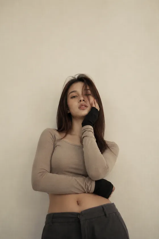 Adult woman with long dark hair posing with crossed arms and one hand at her face in a fitted beige crop top against a plain cream wall.