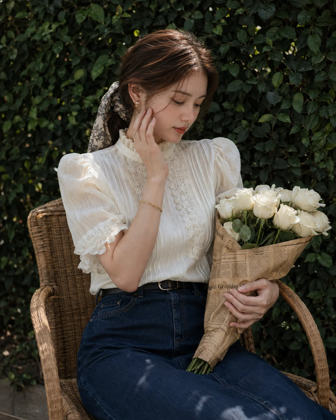 Woman in ivory lace blouse and denim skirt holding white roses on a wicker chair in a garden.