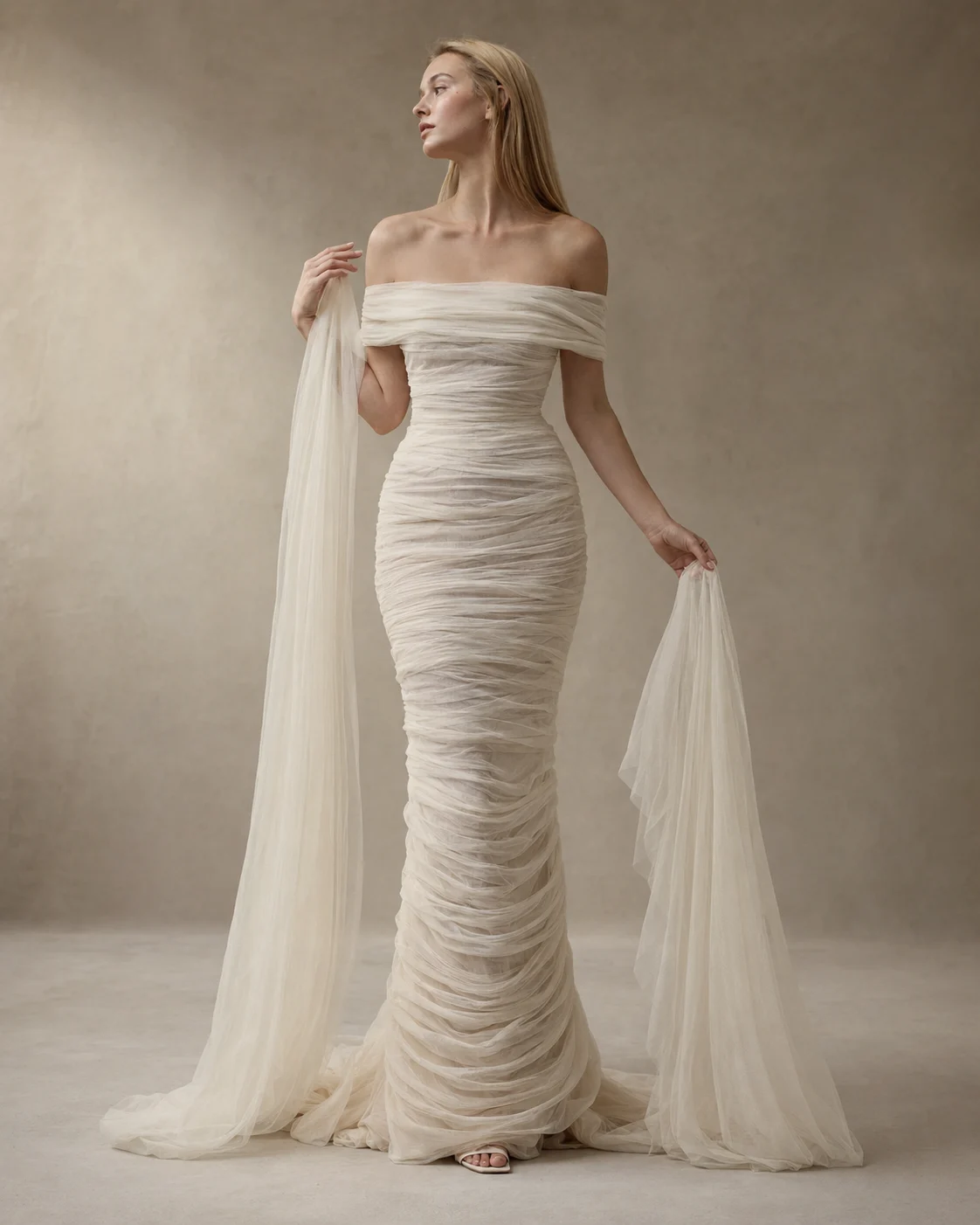 Ethereal ivory off-shoulder ruched bridal gown with long flowing drapes in a minimal studio setting.