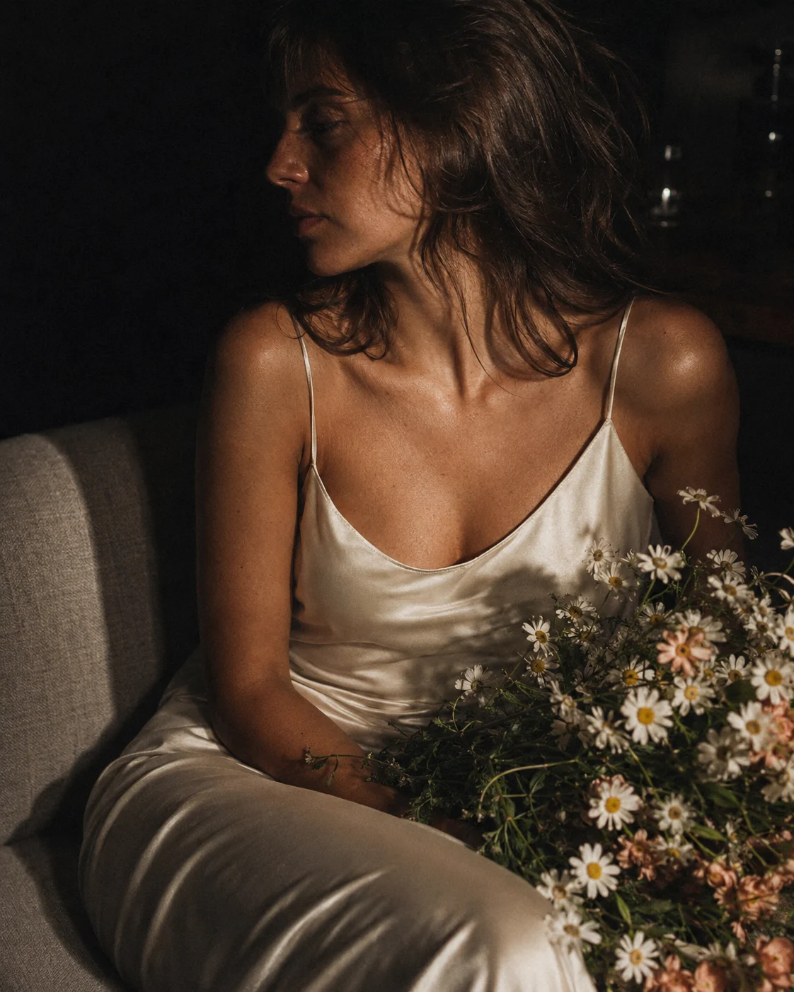 Moody bridal-inspired portrait of a seated woman in an ivory satin slip dress holding white daisies in grainy warm low light.