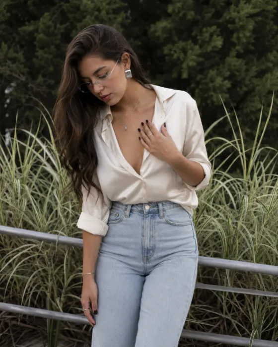 Outdoor fashion portrait with an ivory shirt, light denim jeans, slim glasses, silver jewelry and a cool botanical background.
