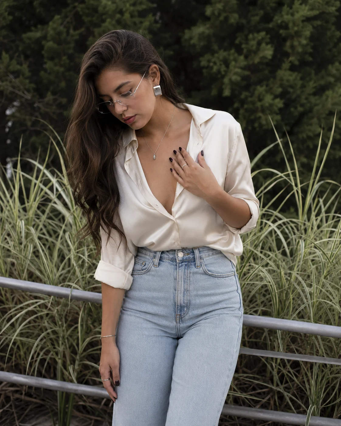 Outdoor fashion portrait with an ivory shirt, light denim jeans, slim glasses, silver jewelry and a cool botanical background.