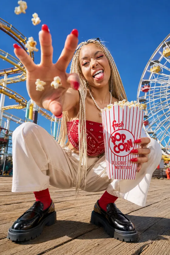 Low-angle amusement park editorial fashion portrait of a subject crouching on a boardwalk with popcorn and rides.