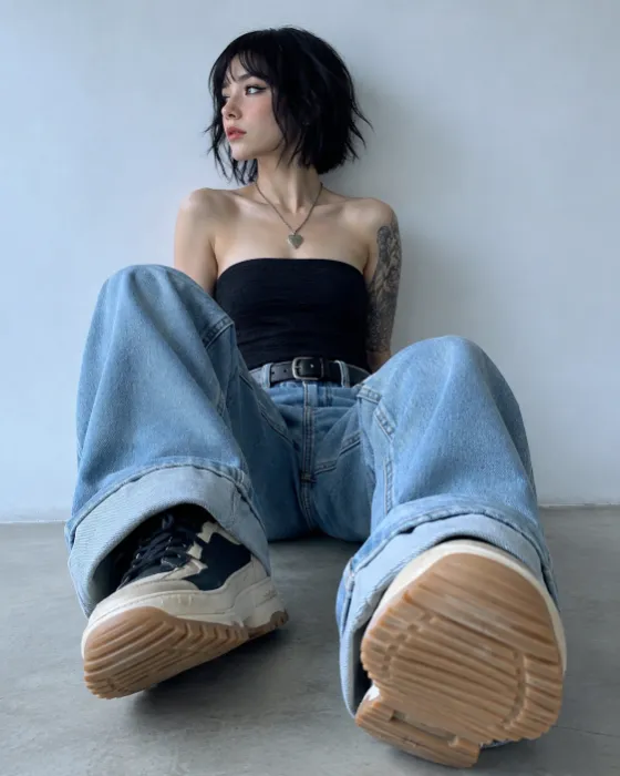 Low-angle seated fashion portrait with oversized blue jeans, black top, and large chunky sneaker soles in the foreground.