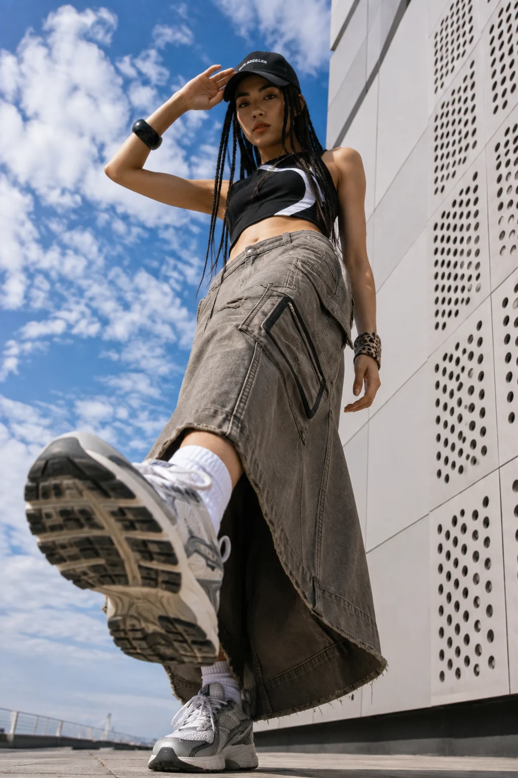 Low-angle urban fashion portrait of a woman in a striped black crop top, cargo maxi skirt, cap, white socks, and chunky sneakers.