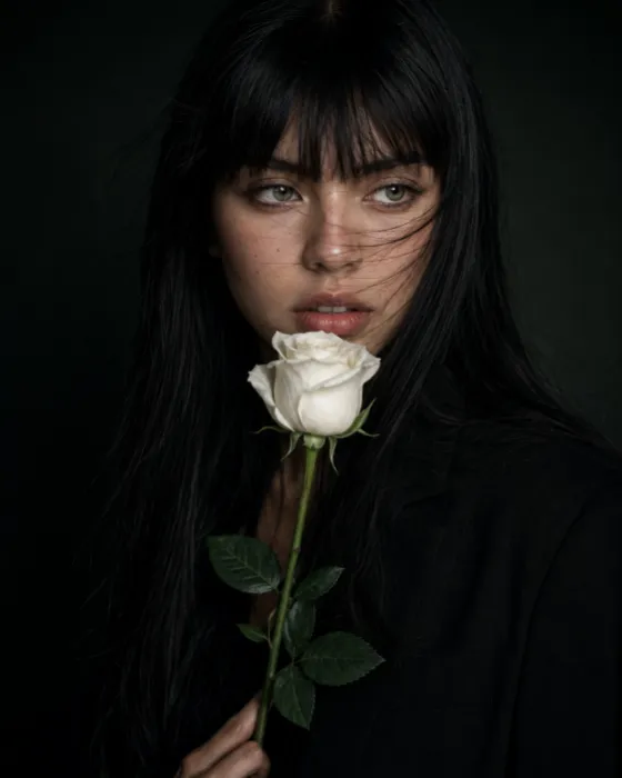 Dark close-up portrait with long black hair, a white rose near the mouth and a softly lit face against a nearly black background.