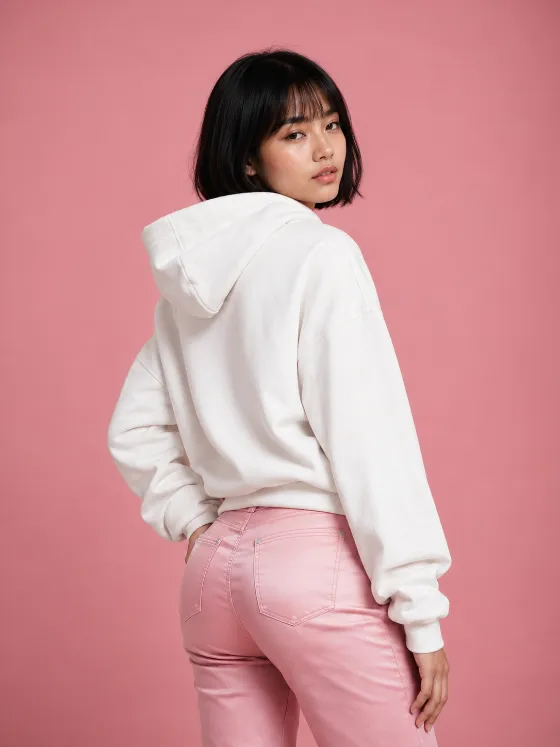 Over-the-shoulder studio fashion portrait with a white hoodie, fitted pink jeans, black bob haircut and matte pink background.