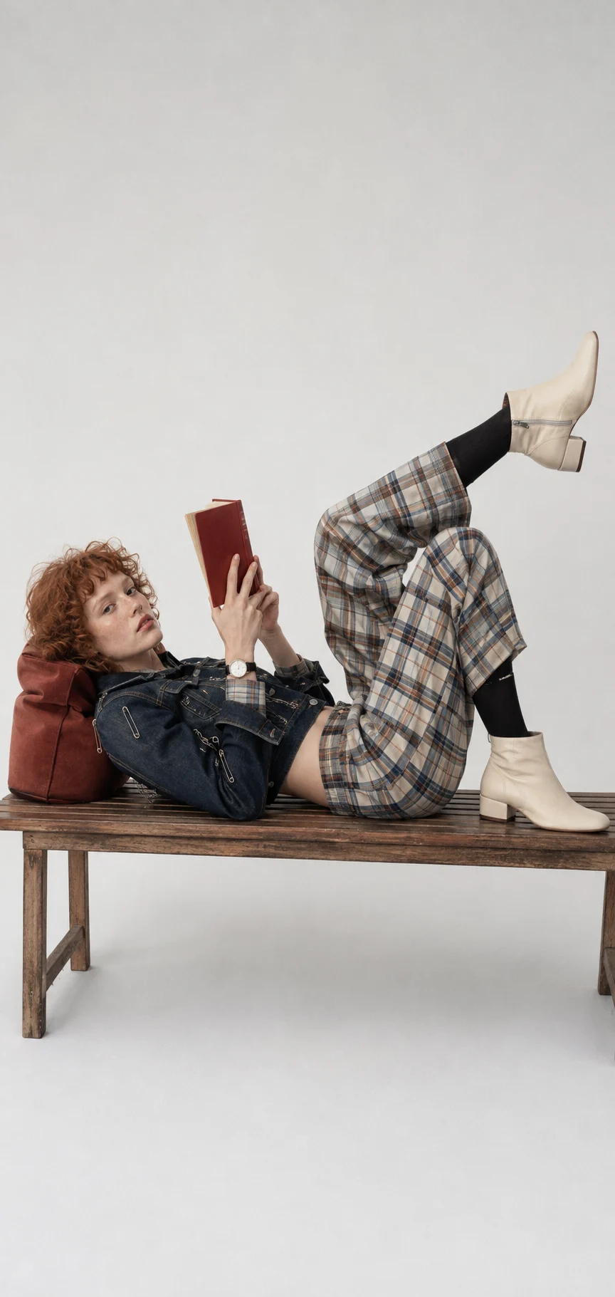 Freckled red-haired woman reclines across a wooden bench in layered denim and plaid, holding a red book with one boot raised.