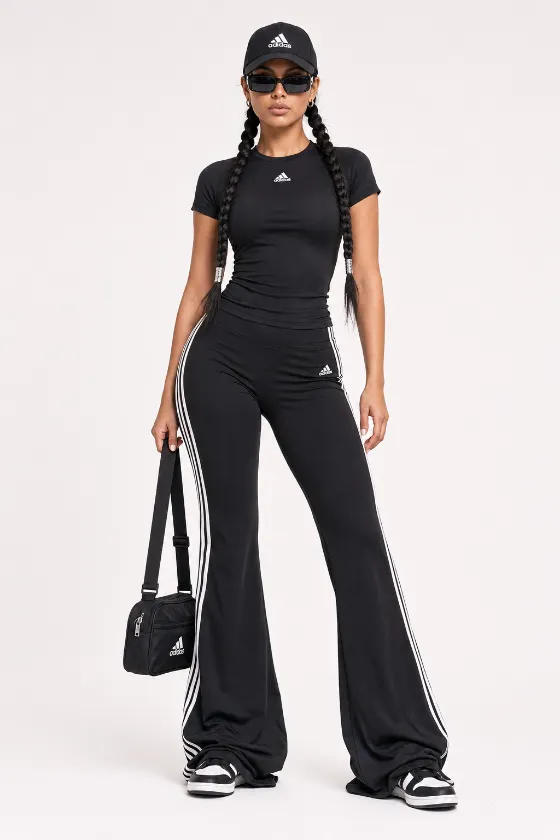 Black monochrome athleisure outfit with flared striped track pants, fitted top, sunglasses, cap, twin braids, and sneakers in a studio setting.