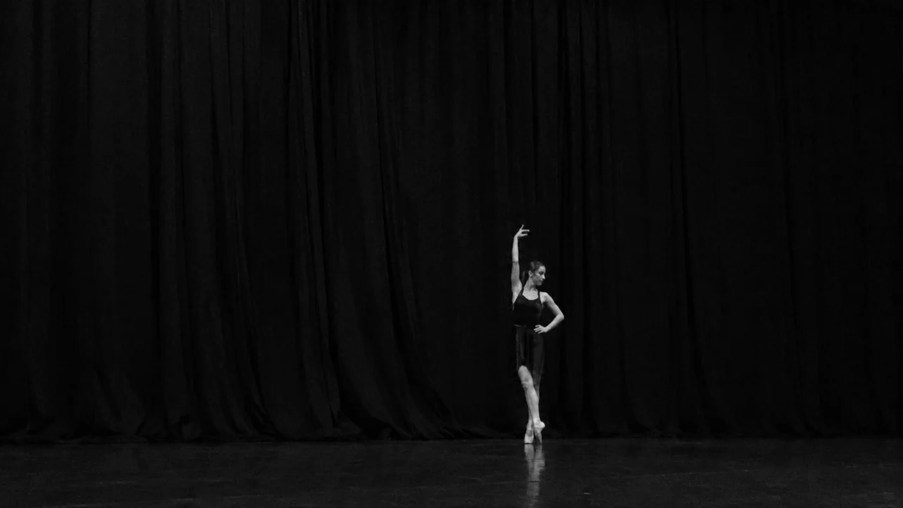 A distant monochrome smartphone-style stage photo of a solitary young ballet dancer posing under dim light in front of a dark draped curtain.