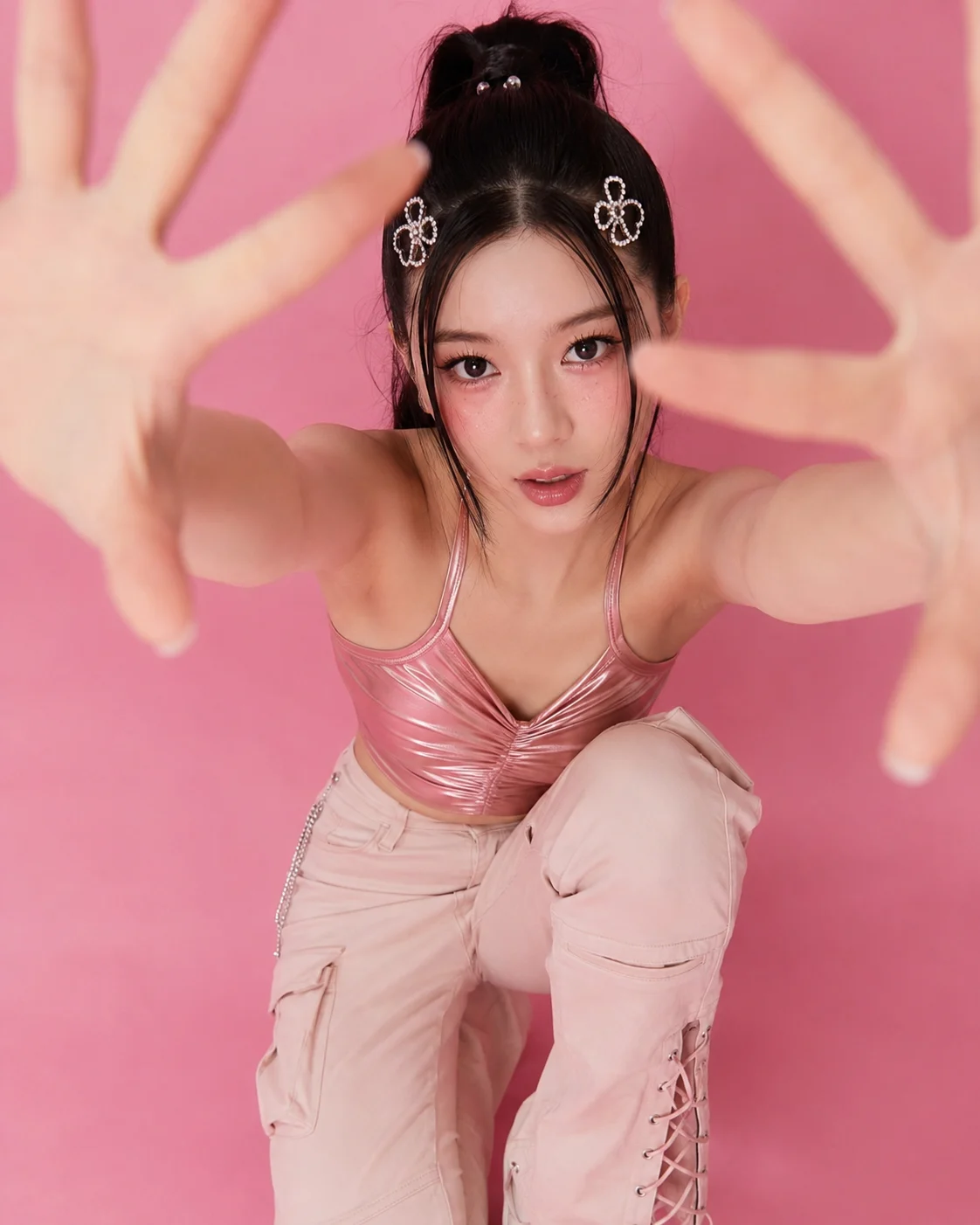 Woman with a high black ponytail and rhinestone clips crouches in pink fashion while reaching both open hands toward an overhead camera.