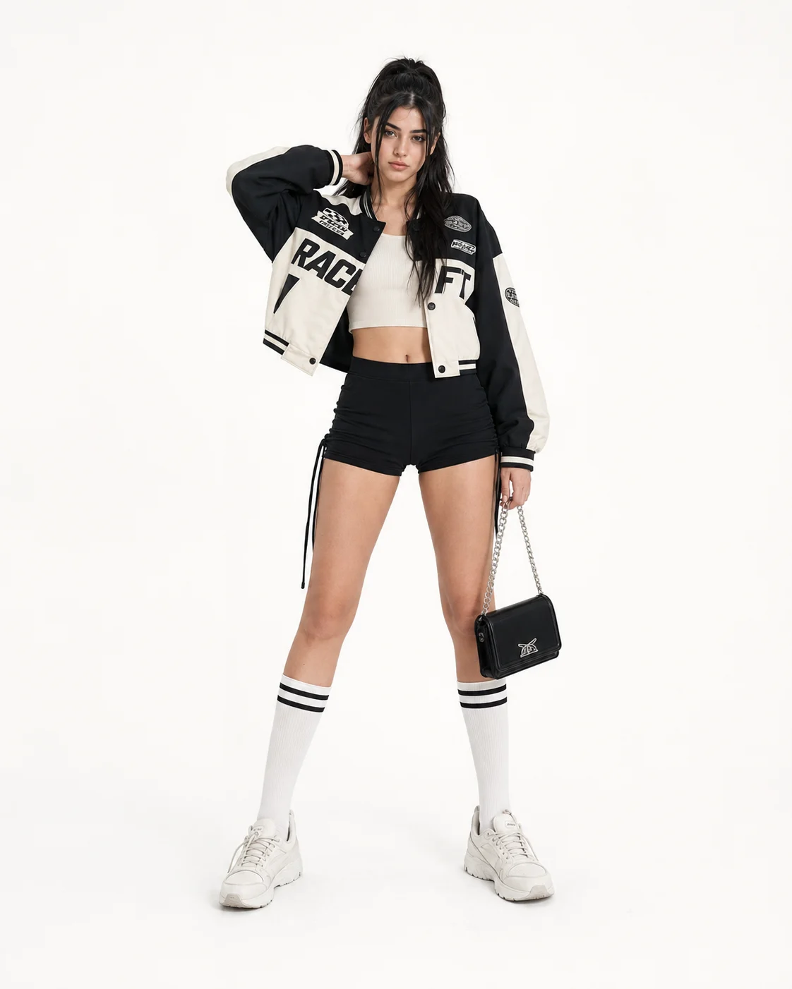 Woman posing frontally in monochrome varsity streetwear with knee socks, chunky sneakers and a black mini bag against a white studio background.